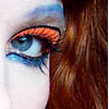 A close-up of a person's eye with stylized makeup, featuring orange and blue eyeshadow, brown hair, and a white background.
