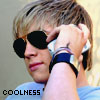 A person with light brown hair wearing dark sunglasses and a light blue shirt holds a silver flip phone to their ear with a hand adorned by multiple wristbands.