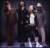 Pixelated static image of multiple figures in dark clothing against a dark background with light highlights on faces and shoulders.