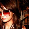 Static close-up of a person with long brown hair wearing large red-tinted sunglasses and layered necklaces, set against an indistinct background.