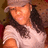 Pixelated image of a person with long dark hair wearing a light-colored cap and white shirt, smiling against a brown background.