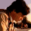 A person with dark hair and light skin wears a light-colored top with a patterned collar, shown in four of a pixelated animation