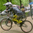A person in a silver helmet and gray jacket rides a yellow bicycle on a dirt path with green foliage and blurred figures in the background.