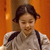 A pixelated image of a person with dark hair tied back, wearing a light-colored shirt with dark straps over the shoulders, against an orange background.