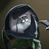 Pixelated figure with dark hooded cloak, light-colored face, and greenish limbs against muted brown background.
