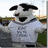 A pixelated image of a cow mascot wearing a gray shirt with text, standing outdoors on grass with buildings in the background.