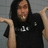 Pixelated image of a person with long dark hair wearing a black shirt with white symbols, arms raised against a gray background.