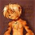 Pixelated figure with light brown hair and orange-toned skin against a dark brown background, wearing a dark red collar and dark brown straps over a light orange torso.
