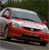 Red sedan with silver roof and white stripe on hood, positioned on road with green background, static image.