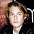 Pixelated close-up of a person with light brown hair wearing a dark jacket over a light shirt against a dark background with a reddish-brown patterned area on the left.
