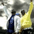 Three figures stand side-by-side against a cloudy sky; the central figure in yellow raises one arm while two others wear blue and white jackets.