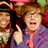Pixelated static image of a character with curly brown hair wearing a purple jacket over a pink shirt, making a middle finger gesture against a dark background with colorful blurred elements.