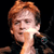 Pixelated close-up of a person with light brown hair holding a microphone against a dark background.