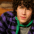 A person with curly brown hair wears a purple and black striped hoodie over a gray shirt with a green drawstring, positioned against a wooden background.