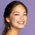 Pixelated profile view of a person with dark hair tied back, light skin, and a smiling expression against a solid purple background.