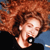 Pixelated image of a person with voluminous curly red hair, wearing a dark top and blue garment, mouth open in a shout or laugh.