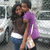 Two people embrace in front of parked cars, one wearing a purple shirt and white belt with blue pants.