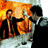 Pixelated static image of two figures in dark clothing against a fiery orange-yellow background with visible dithering and compression artifacts.