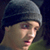 Static pixelated image of a person wearing a dark beanie and light-colored shirt against a dark background with green foliage at the bottom.
