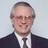 Static pixelated image of a person in formal attire with short graying hair, wearing glasses and a dark suit jacket over a white shirt and striped tie.