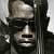 Pixelated grayscale image of a person's face with dark hair and glasses, wearing a dark jacket over a light shirt against a neutral background.