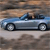 A light blue convertible car with a black soft top is shown in side view, positioned on a light gray road against a brown textured background.