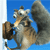 Pixelated cartoon animal with brown and gray fur, white chest, and large eyes, holding a brown object against a solid blue background.