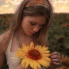 A woman with long light brown hair wears a white halter top and a beaded necklace while holding a large yellow sunflower with a brown center in a field of blurred sunflowers under a soft sky.