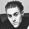 A black-and-white portrait of a person with short curly hair and dark eye makeup, wearing a dark shirt against a light background.