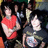Two people in red and black shirts pose with arms crossed against a blurred background of other figures.
