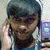 A person with dark hair wearing a blue top holds a purple device near their face, with hand positioned near ear, in a static image with pixelated quality.