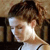 Pixelated close-up of a person with brown hair tied back, wearing a white top with dark straps, against a blurred background.