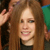 A pixelated close-up of a person with long brown hair wearing a dark top, smiling with visible teeth, and raising a hand near their head.