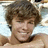 Pixelated close-up of a person with brown curly hair, light skin, and a neutral expression against a light blue and white background.