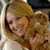 A woman with long wavy blonde hair smiles while holding a small brown dog, wearing a light-colored hooded garment.