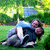 Two people embrace on grass under trees, one in blue shirt and dark pants, the other in light top and white pants.