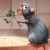 A gray cartoon rat with orange ears and nose holds a green leafy sprig in a kitchen setting with brown walls and a light counter.