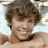 A person with curly brown hair lies on a light blue patterned surface, smiling broadly in three showing slight head movement