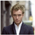Pixelated image of a person with light brown hair wearing a dark suit jacket over a white shirt, standing against a blurred background with vertical and horizontal elements.