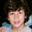 A pixelated image of a person with curly brown hair and light skin, wearing a white shirt against a dark background.