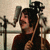 Pixelated image of a person with dark hair holding a stringed instrument, standing before microphones in an indoor setting.