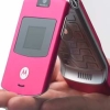A pink flip phone held in a hand, showing its front screen and open internal keypad with a patterned surface.