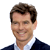 Static pixelated image of a smiling man with brown hair wearing a dark suit jacket over a white shirt against a plain background.