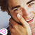 Two people in close proximity, one covering the other's eyes with a hand, both smiling, with a pink and white pixelated symbol in the bottom left corner.