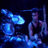 A drummer in a black tank top plays drums under blue lighting with cymbals and drum kits visible.