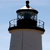 A pixelated static image of a lighthouse tower with a black lantern room, white trim, and brown base against a light blue sky.