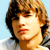 Pixelated static image of a person with light brown hair and fair skin against a light blue background, wearing a white shirt with dark stripes on the shoulder.
