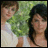 Two people stand side-by-side against a green leafy background, one in white clothing with brown hair and the other in light-colored attire with dark hair.