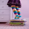 A child's legs in rainbow striped tights and dark skirt stand on a stack of books near a white dresser.