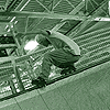 A person in a crouched position on a skateboard under green metal structures, transitioning to standing and riding down stairs with railings.